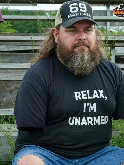 Man med skägg, keps och T-shirt med texten "Relax, I'm unarmed" sitter på träbänkar utomhus.