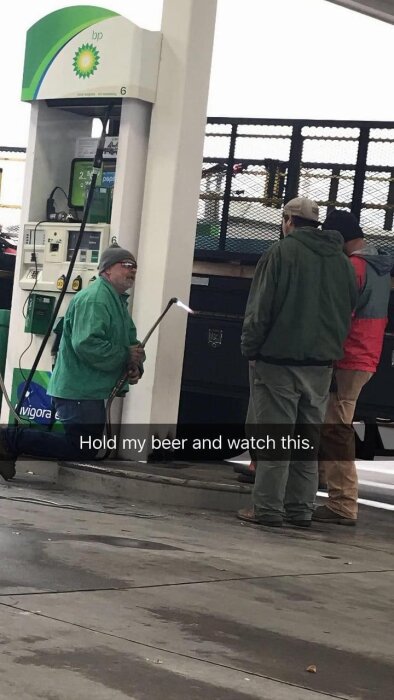 Tre män vid en bensinpump medan en av dem håller en tänd svets. Bensinpumpen tillhör BP. Textruta i bilden lyder: "Hold my beer and watch this.