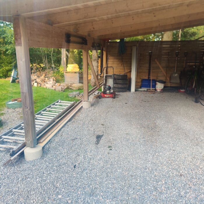 Bild på träram med tak över grusbelagd carportihopkopplad med garage, stege liggande på marken, däck och redskap lutade mot garageväggen i bakgrunden.