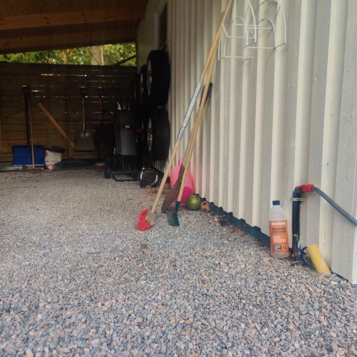 Carport med grusgolv, verktyg och redskap mot garagevägg inklusive borstar och hinkar, och en sprayflaska vid väggen.