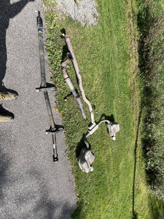 Borttagna bildelar, inklusive avgasrör och andra metalldelar, ligger på en uppfart vid gräs. Skugga av en person ses på trottoaren.