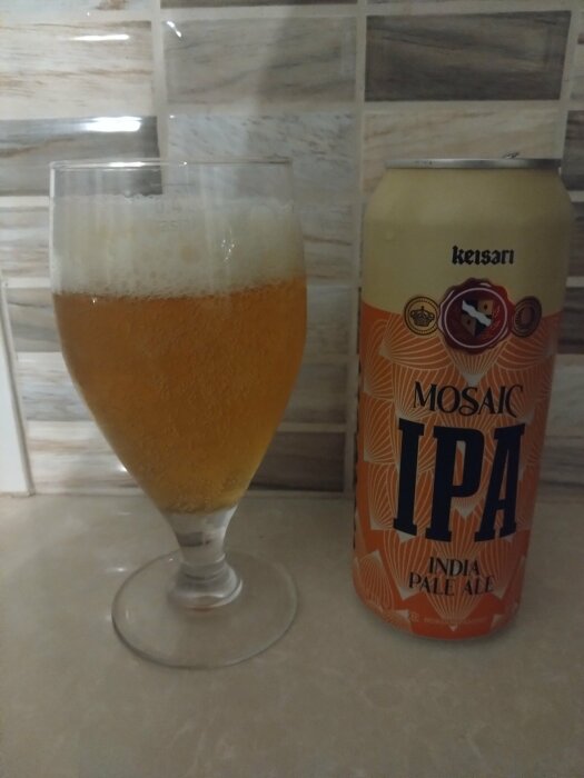 En burk Keisari Mosaic IPA India Pale Ale och ett glas fyllt med samma öl framför ett kaklat vägg.