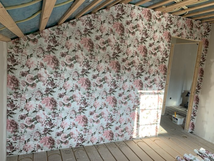 Blommig tapet med rosa och lila blommor på en vägg i ett rum under renovering med synliga takbjälkar. Door ram utan dörr och oavslutat trägolv.