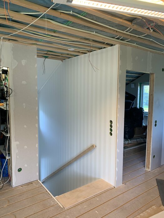 Renovering av övervåning med exponerat tak och elinstallationer, tapetserad vägg i trapphus, synlig isolering och sladdar, träbjälkar synliga i taket.