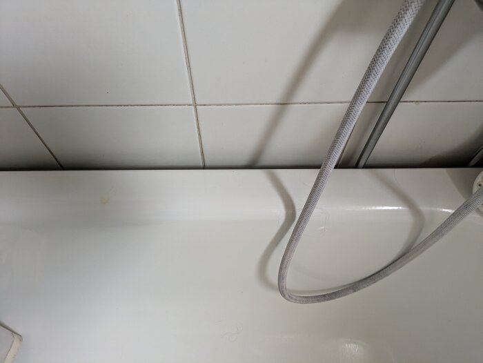 Glapp på 2,5-3 cm mellan badkar och kaklad vägg med grå slang i högra hörnet.