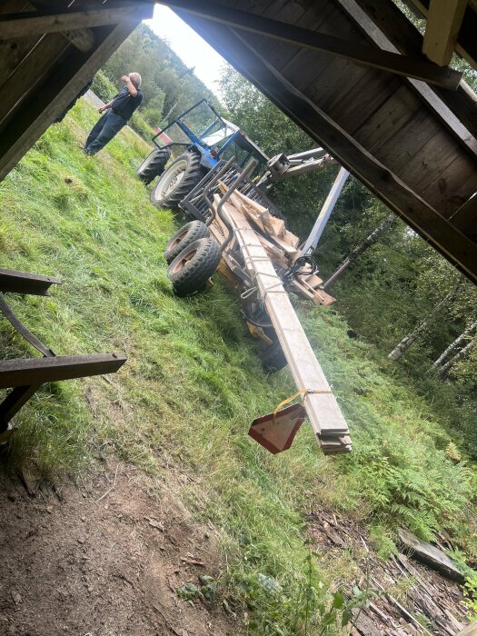 Traktor med släp lastat med långa träplankor på en gräsbevuxen tomt, en man står i närheten och skymtar i bildens vänstra hörn.