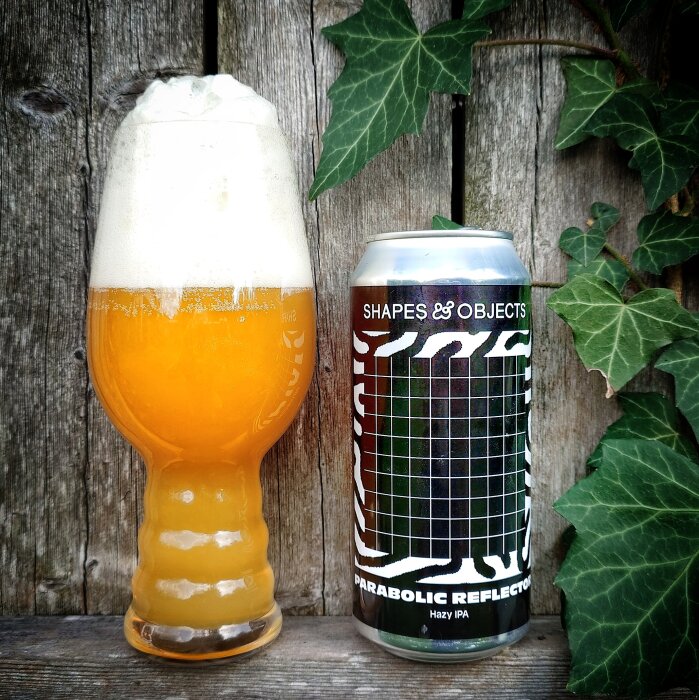 En glas med skummande hazy IPA ligger bredvid en burk Shapes & Objects Parabolic Reflector Hazy IPA framför en trävägg med murgröna.