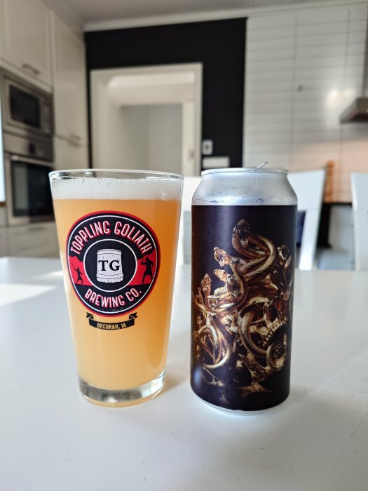 Ett ölglas med en orange öl från Toppling Goliath Brewing Co. bredvid en burk med mörk konstnärlig design på ett bord i ett modernt kök.