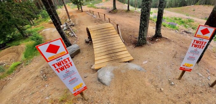 Mountainbikestig i skogen med en träbro och skyltar som markerar "LöpLed T6 TWIST TWIST" för avancerad nivå placerad på båda sidor av bron.