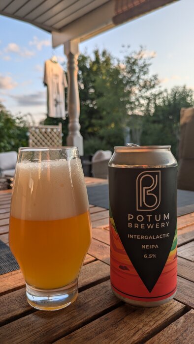 Ett glas med skummande öl och en burk Potum Brewery Intergalactic NEIPA, 6,5% på ett träbord ute på en veranda.