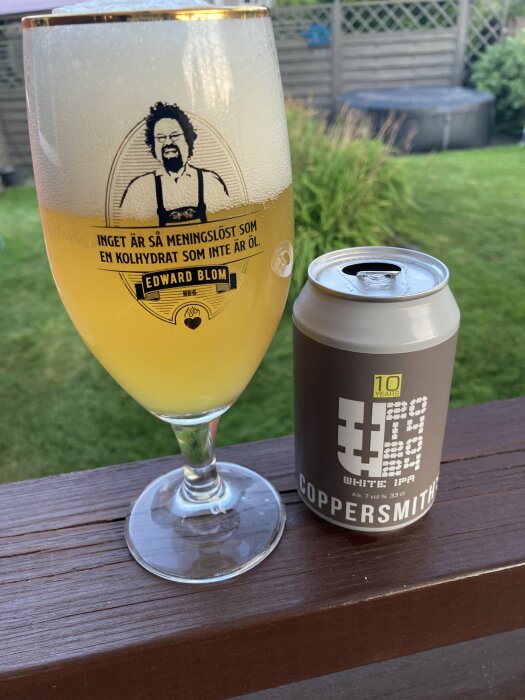 Ölglas med text och bild av Edward Blom står bredvid en burk Coppersmith White IPA på ett staket vid en gräsmatta.