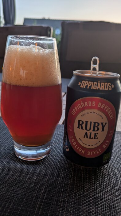 Ett glas med skummande röd ale bredvid en öppnad burk Oppigårds Ruby Ale på ett bord utomhus, med stolar i bakgrunden.