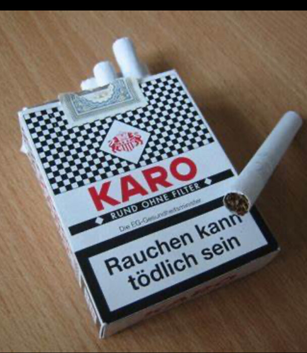 En öppnad cigarettask med texten "KARO" och "Rauchen kann tödlich sein" ligger på ett träbord, med cigaretter synliga.