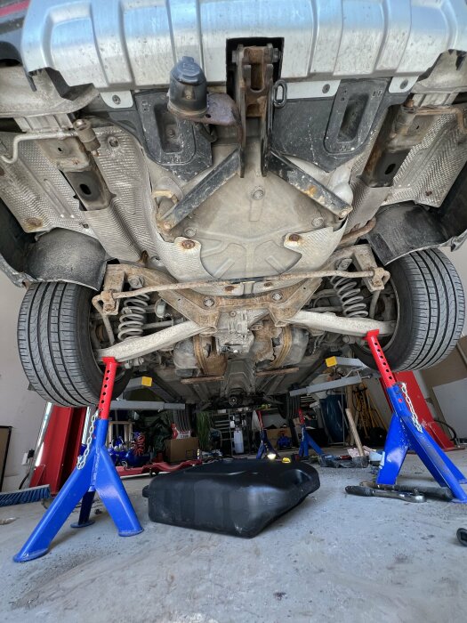 Bil lyft på pallbockar med bränsletank undertill i garage, verktyg spridda på golvet.