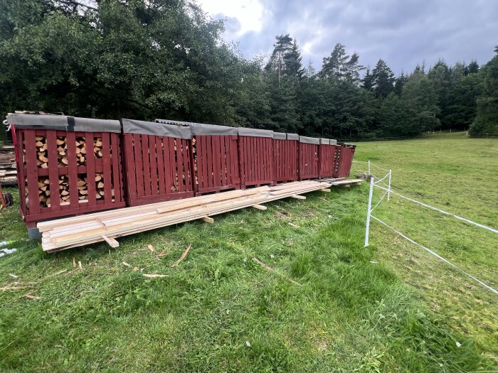 Hög med torkande virke och ved i röda träställningar på en gräsmatta, med träd och skog i bakgrunden under en mulen himmel.