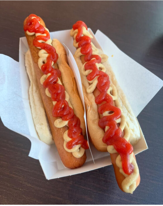 Två korv med bröd, en kokt och en grillad, toppade med senap och ketchup, serverade i en pappersbehållare på en mörk bordsyta.