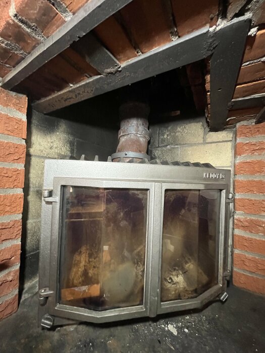 Insatsen i en öppen spis, omgiven av tegel och metall, med ett rör ovanpå och spår av sot på glasdörrarna.