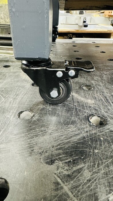 Närbild av ett nytt svart låsbart hjul monterat på en grå hyvelmaskin, stående på en repig arbetsyta av metall.