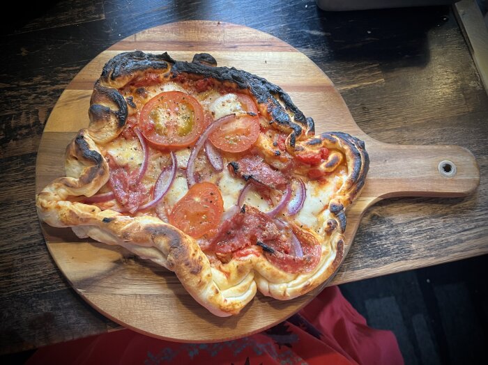 Hemlagad pizza med tomatsås, mozzarella, rödlök, salami och färska tomater, något ojämnt gräddad och brunad på vissa områden, på en pizzaspade.