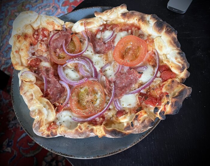 Hemmagrillad pizza med ost, tomatsås, mozzarella, rödlök, salami och färska tomater, delvis undergräddad och brandskadad på kanterna.