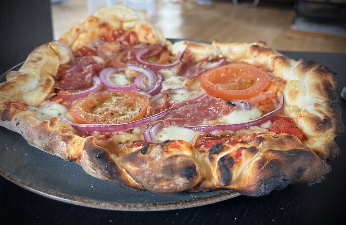 Hemlagad pizza med tomatsås, mozzarella, rödlök, salami, färska tomater och ost på en tallrik, något bränd i kanterna.