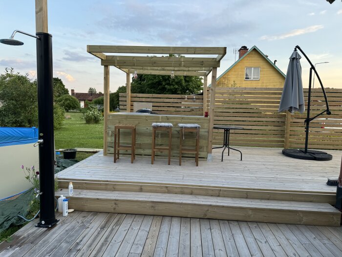 Trädäck med ett utomhuskök, pergola och barstolar. Trädgårdsdusch till vänster och trädgårdsmöbler med parasoll till höger. Gul villa i bakgrunden.