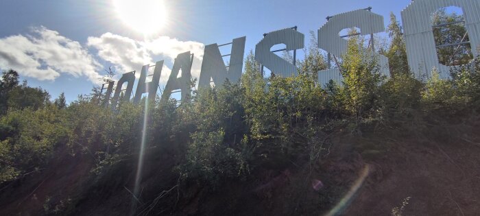 Stora vita metallbokstäver som bildar ord i ett skogsområde, med solen och blå himmel i bakgrunden.