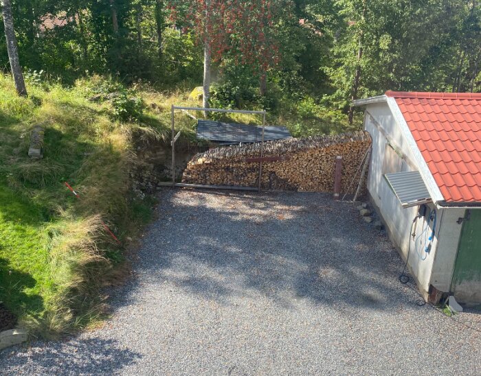 Grusplan framför ett garage med rött tegeltak. Till vänster finns en sluttning med gräs och en vedstapel bakom en metallram. Skog i bakgrunden.