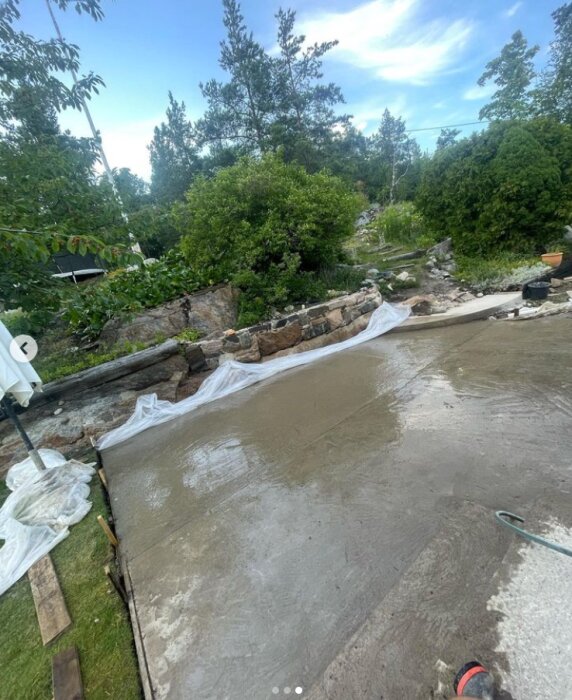 Nyutlagd betongplatta i en trädgård, omgiven av stenar och grönska. Blicken riktad mot en stenmursomringad yta som väntar på att torka och härda.