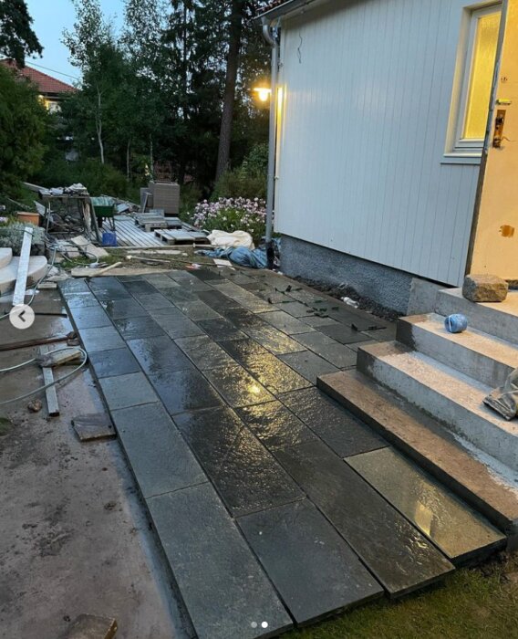 Nyinstallerad skifferbeläggning framför ett hus med vita väggar, trappa till entrén och diverse byggmaterial spritt runtom området.