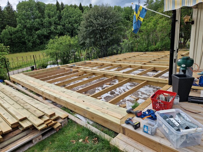 Påbörjat byggprojekt av veranda, med träbjälklag färdigställt och trallvirke redo att läggas på. Verktyg och byggmaterial ligger framme, och en svensk flagga hänger i bakgrunden.