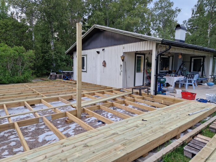 Ramverket av en veranda under konstruktion vid ett hus, med ett delvis färdigt golv av trallbrädor och verktyg spridda omkring.