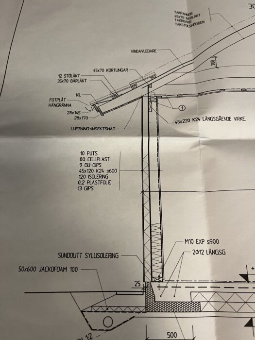 Ritning av husets konstruktion som visar detaljer som isolering, gipsskivor, och syllisolering. Vinden, takfoten och sockeln är utmärkta med mätningar.