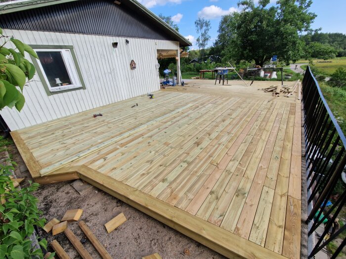 Nybyggd veranda med trägolv till ett vitt hus, arbetsredskap ligger utspridda och träräcken monterade längs verandans kant.