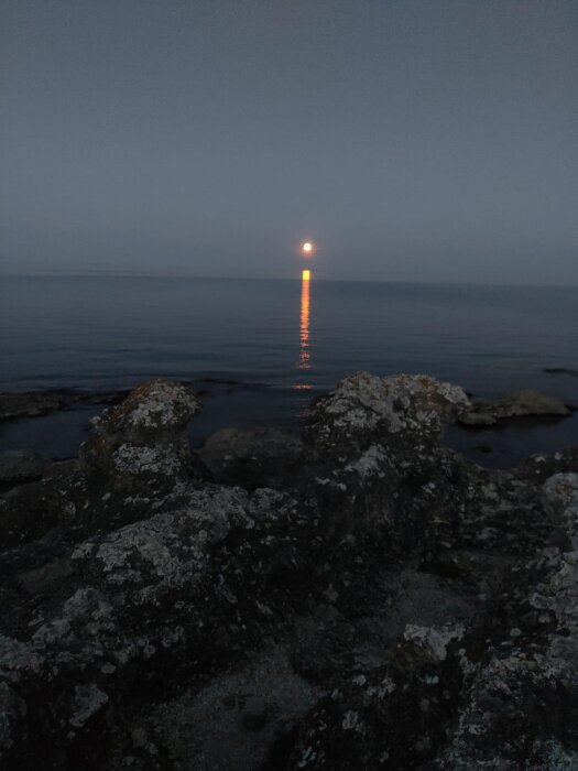 Solnedgång över ett lugnt hav, sedd från en klippig kustlinje. Solens reflektion sträcker sig som en ljusstråle från horisonten till klipporna i förgrunden.