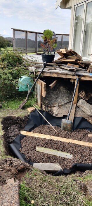 Grävd grop med lecakulor för badtunna vid altan, verktyg och jordhögar synliga, temporär avlägsnad trappa och växtkrukor på altanen.