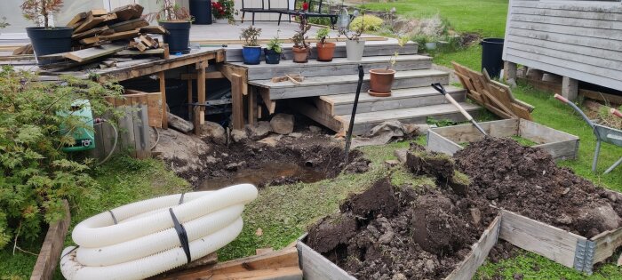 Borttagen altantrappa med flera blomkrukor ovanpå. En stor grop fylld med vatten, grushögar och dräneringsrör synliga på marken, samt byggverktyg runt omkring.