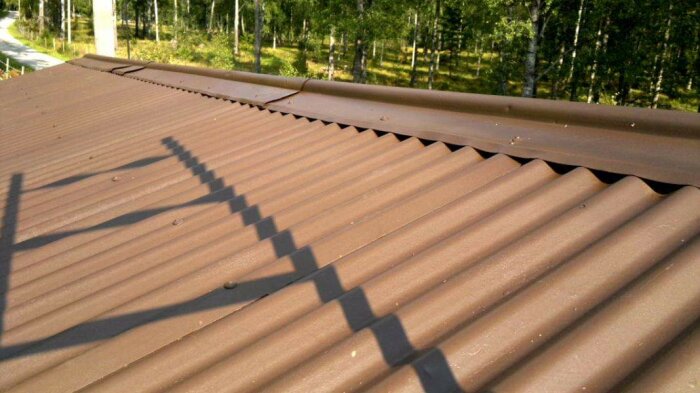 Brunt, korrugerat tak fotograferat i solen med skuggor från en stege. Bakgrunden består av skogsmiljö med grusvägar.