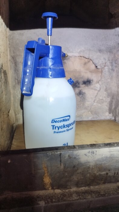 En plastbehållare från DecoMax med texten "Tryckspruta Pressure Sprayer" står inuti en vittrad eldstad med synliga skador och beläggningar på insidan.