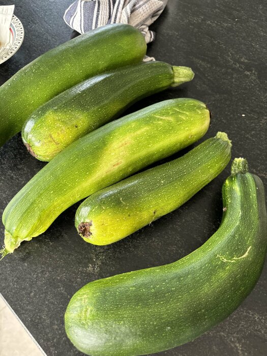 Fem zucchini ligger på ett mörkt köksbänkskiva, med en kökshandduk i bakgrunden.