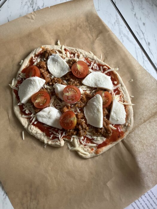 En oregelbundet formad pizza på bakplåtspapper med tomatsås, riven ost, körsbärstomater, köttfärs och skivor av mozzarellaost, redo att gräddas.