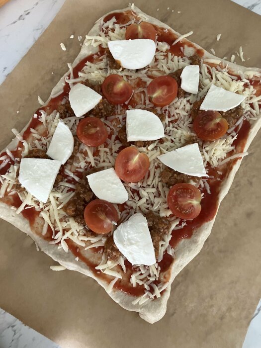 En rektangulär pizza med tomatsås, riven ost, skivade körsbärstomater och bitar av mozzarella på bakplåtspapper, redo för pizzasten.