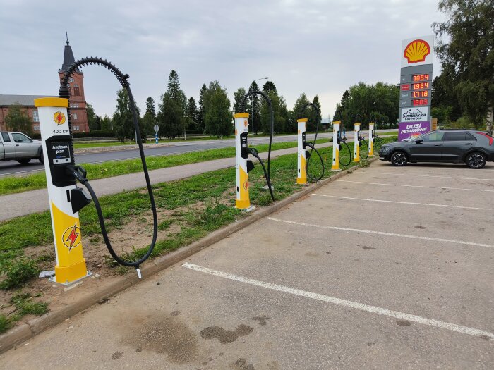 Sex laddstationer för elbilar med skylten "Avataan pian" på en finsk bensinmack. Shell-logotyp och bränslepriser syns i bakgrunden. En SUV står parkerad.