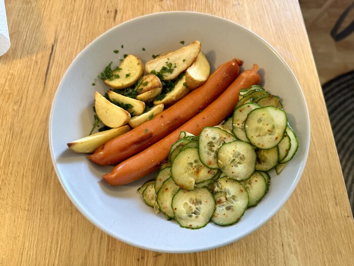 En tallrik med korv, potatis och gurksallad på ett träbord.