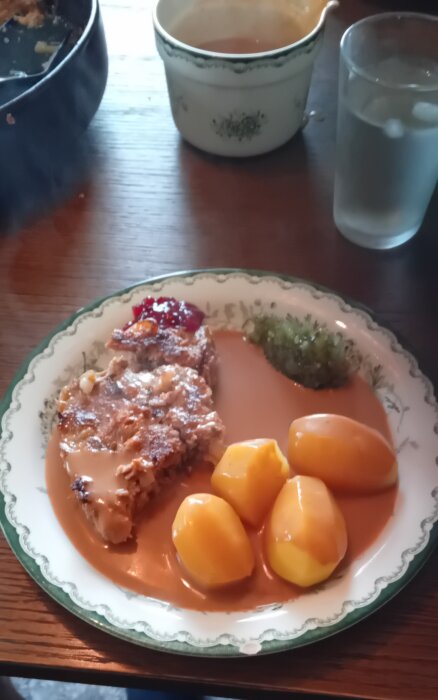 En tallrik med kålpudding serverad med fyra potatisar, sås, lingonsylt och en klick grönsaksröra på ett bord tillsammans med en kopp och ett glas vatten.