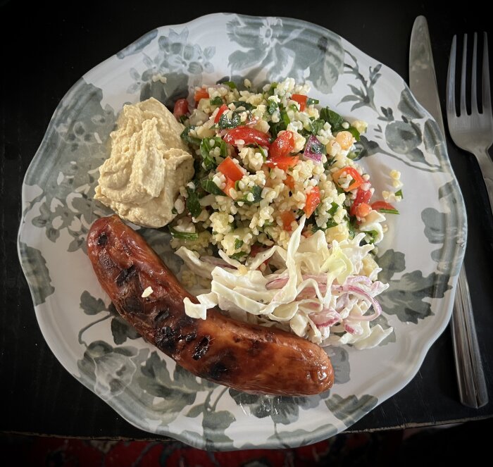 En tallrik med en grillad bratwurst, tabbouleh med paprika, gurka och fetaost, hummus samt coleslaw.