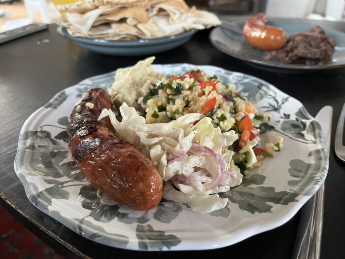 En tallrik med grillad bratwurst, coleslaw, tabbouleh, hummus och libabröd i bakgrunden.