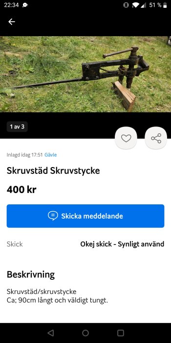 Gammalt skruvstycke liggande på gräs, ca 90 cm långt med en tung metallkonstruktion och träblock.