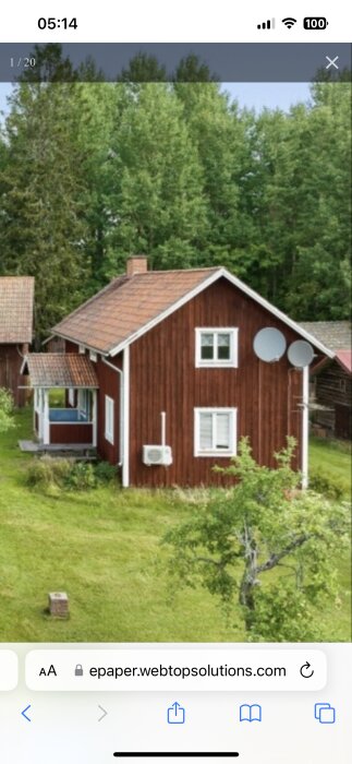 Rött trähus med tegeltak, två fönster, två satellitantenner och en luftkonditioneringsenhet på sidan. Omgiven av grönska och träd i bakgrunden.