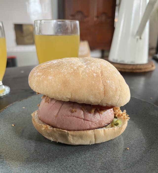 En parisare i surdegsbröd med ketchup, senap, bostongurka och rostad lök på en tallrik, med två glas dryck i bakgrunden.